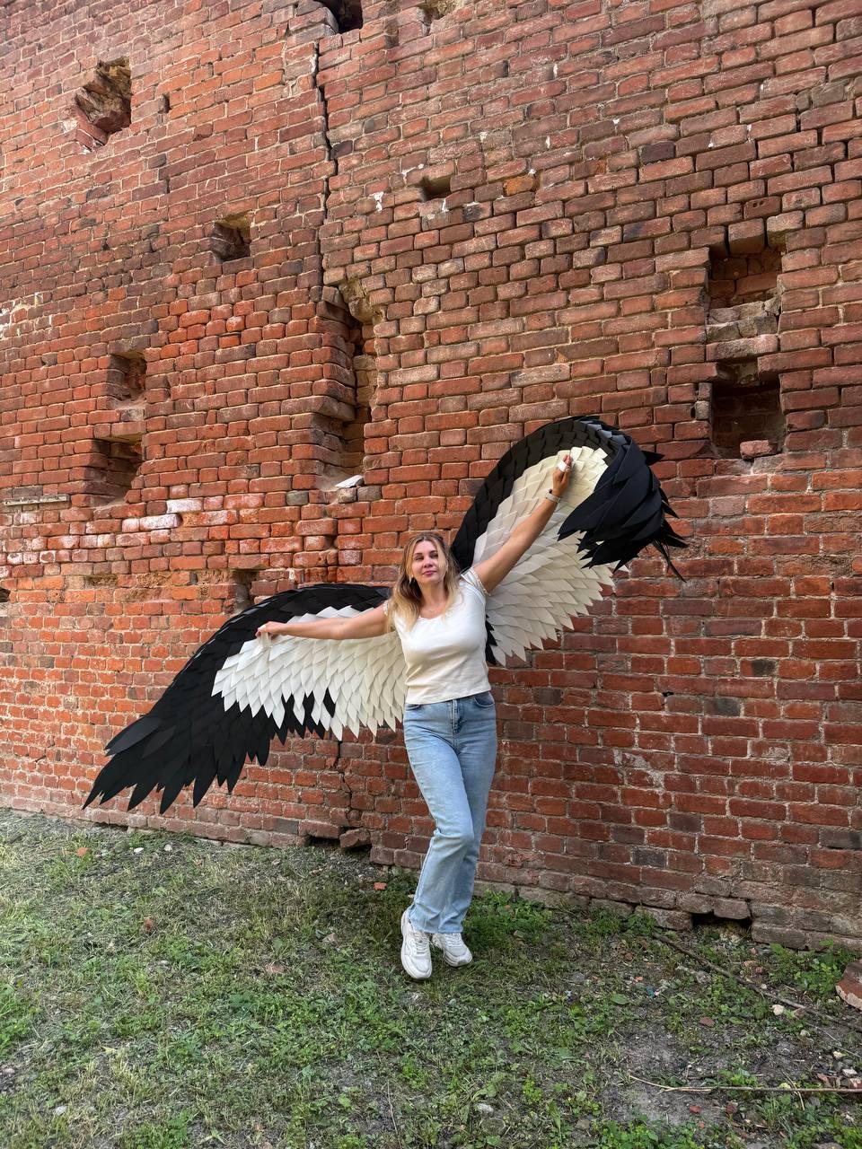 May include: Large wearable wings featuring a layered design with white inner feathers and black outer feathers. The wings are constructed with a rigid frame and extend horizontally, showcasing a sharp, pointed silhouette at the tips of the feathers.