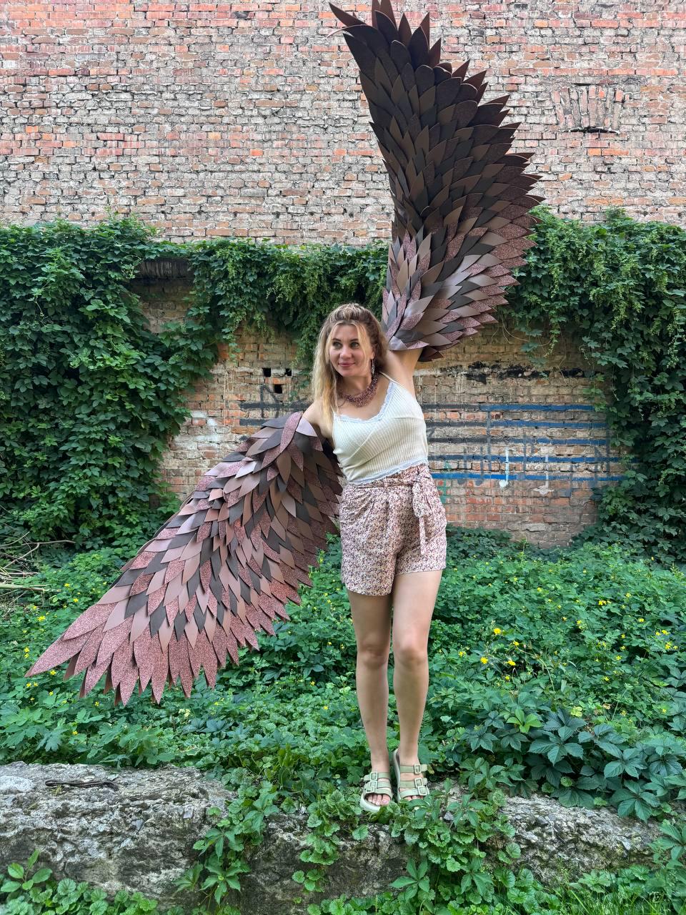 May include: Large, articulated wings constructed from numerous overlapping dark brown and copper-toned feather-shaped pieces. The wings feature a layered, textured design that mimics the appearance of avian plumage, extending outward from a central harness.