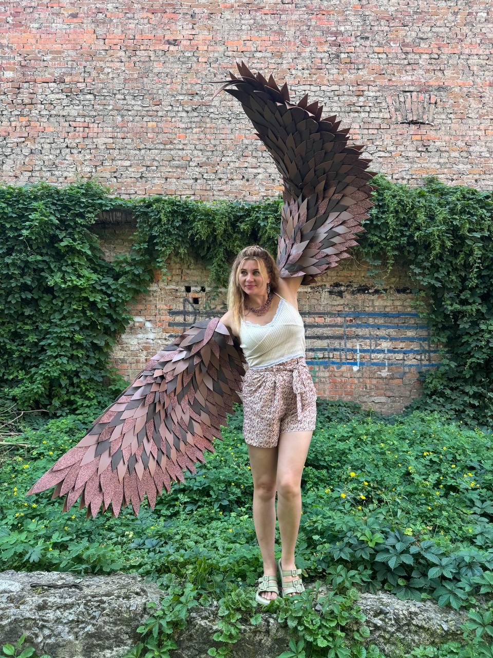 May include: Large, articulated wings constructed from layered, dark brown and copper-toned leaf-shaped feathers. The wings extend outward, featuring a textured, metallic finish that catches the light, creating a dramatic, expansive span of plumage.