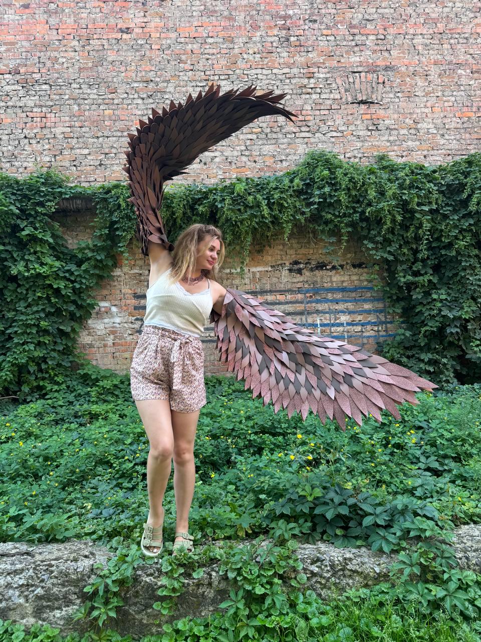 May include: A pair of large, articulated costume wings featuring layered, dark brown, feather-like panels. The wings are designed to be worn on the back, with one wing extended upward and the other extended horizontally to display the detailed structure.