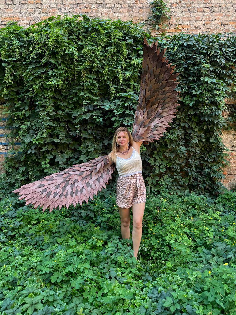 May include: A pair of large, articulated costume wings featuring layered, feather-like segments in shades of brown and tan. The wings are designed to be worn on the back, extending outward to create a wide, expansive span with a textured, organic appearance.