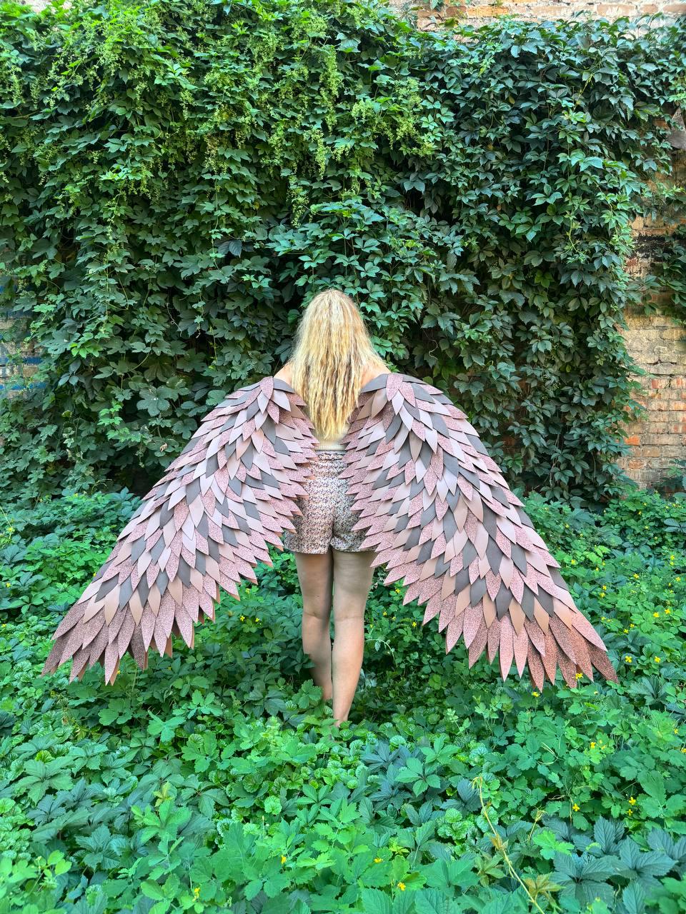 May include: Large decorative wings featuring layered, pointed feathers in shades of dusty rose, mauve, and dark brown. The wings are constructed with a textured, matte finish and are designed to be worn on the back, extending outward in a wide span.