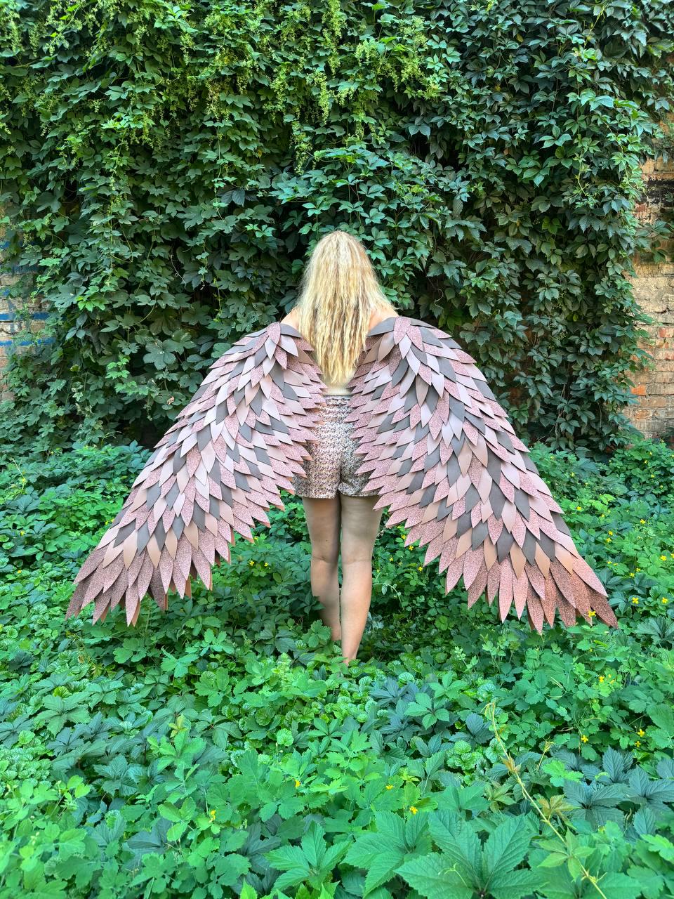 May include: A pair of large, wearable wings featuring layered, leaf-shaped feathers in shades of muted pink, mauve, and dark brown. The wings are constructed with a textured, glittery finish on the individual feather segments, creating a dense, overlapping pattern that spans outward from the center.