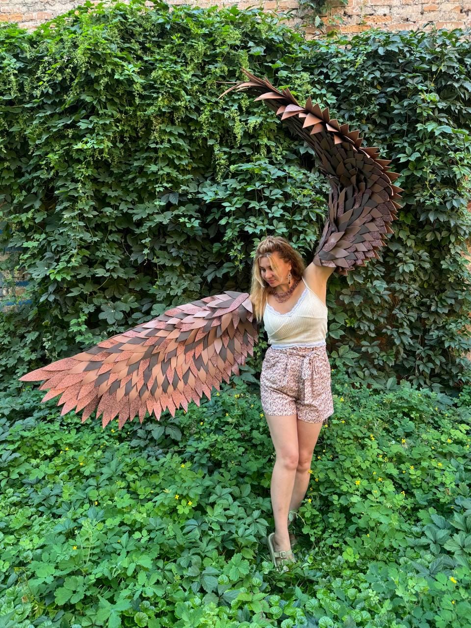 May include: A pair of large, articulated wings constructed from layered, leaf-shaped pieces in shades of brown and copper. The wings feature a textured, feathered appearance with a wide span, designed to be worn on the back with visible straps.