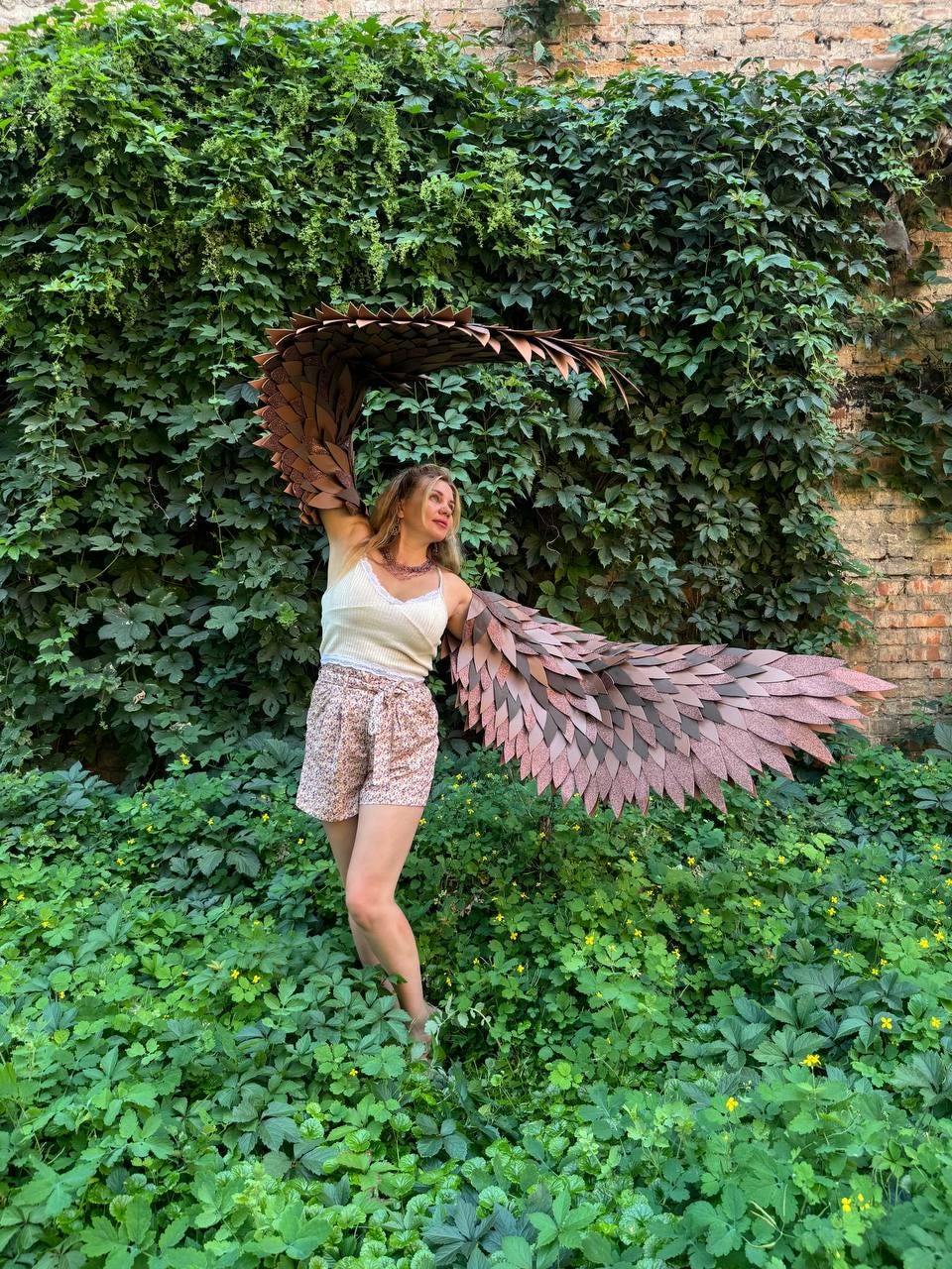 May include: A pair of large, articulated brown wings with layered, feather-like segments attached to a white sleeveless top and patterned shorts. The wings extend outward, showcasing a textured, rigid construction with a dark brown finish.