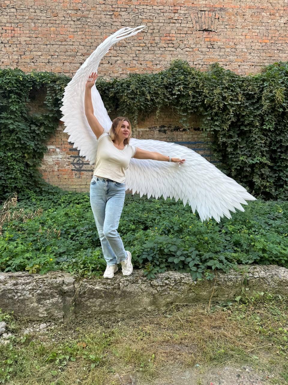 May include: A pair of large, white, feathered-style wings attached to a beige short-sleeved t-shirt and light blue denim jeans. The wings feature a layered, pointed design that extends outward, with one wing reaching upward and the other extending horizontally.