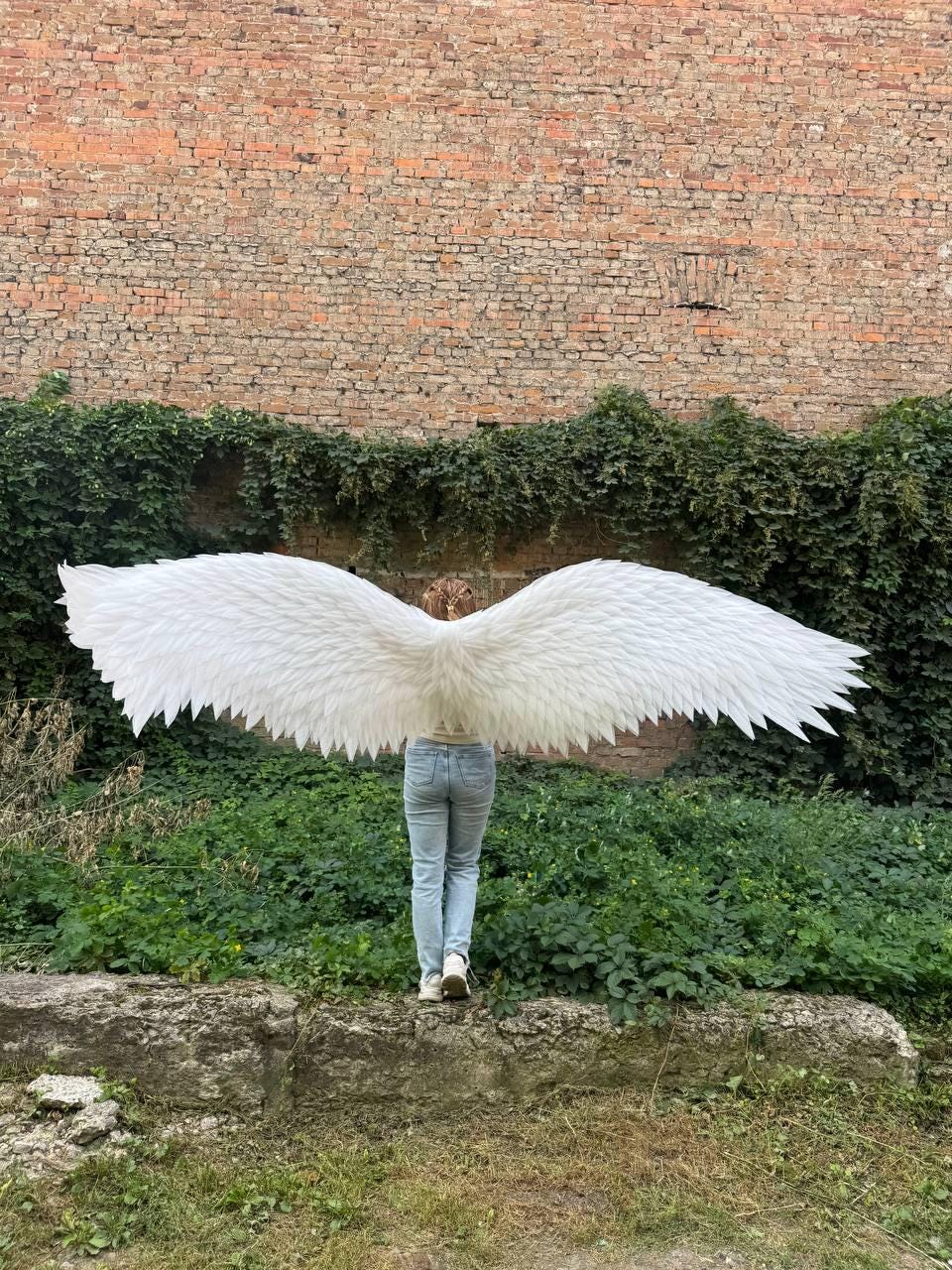 May include: A pair of large, white, feathered wings attached to a harness, worn over light blue denim jeans. The wings are fully extended horizontally, showcasing detailed, layered white plumage that spans a wide width across the frame.
