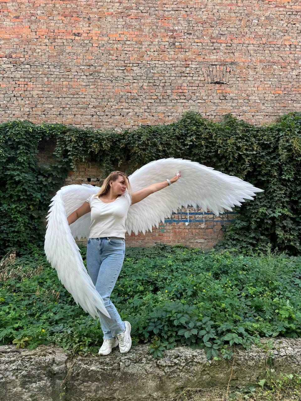 May include: Large, white, feathered angel wings attached to a harness worn over a white short-sleeved t-shirt, light blue denim jeans, and white sneakers. The wings feature layered, pointed white feathers that extend outward to both sides.