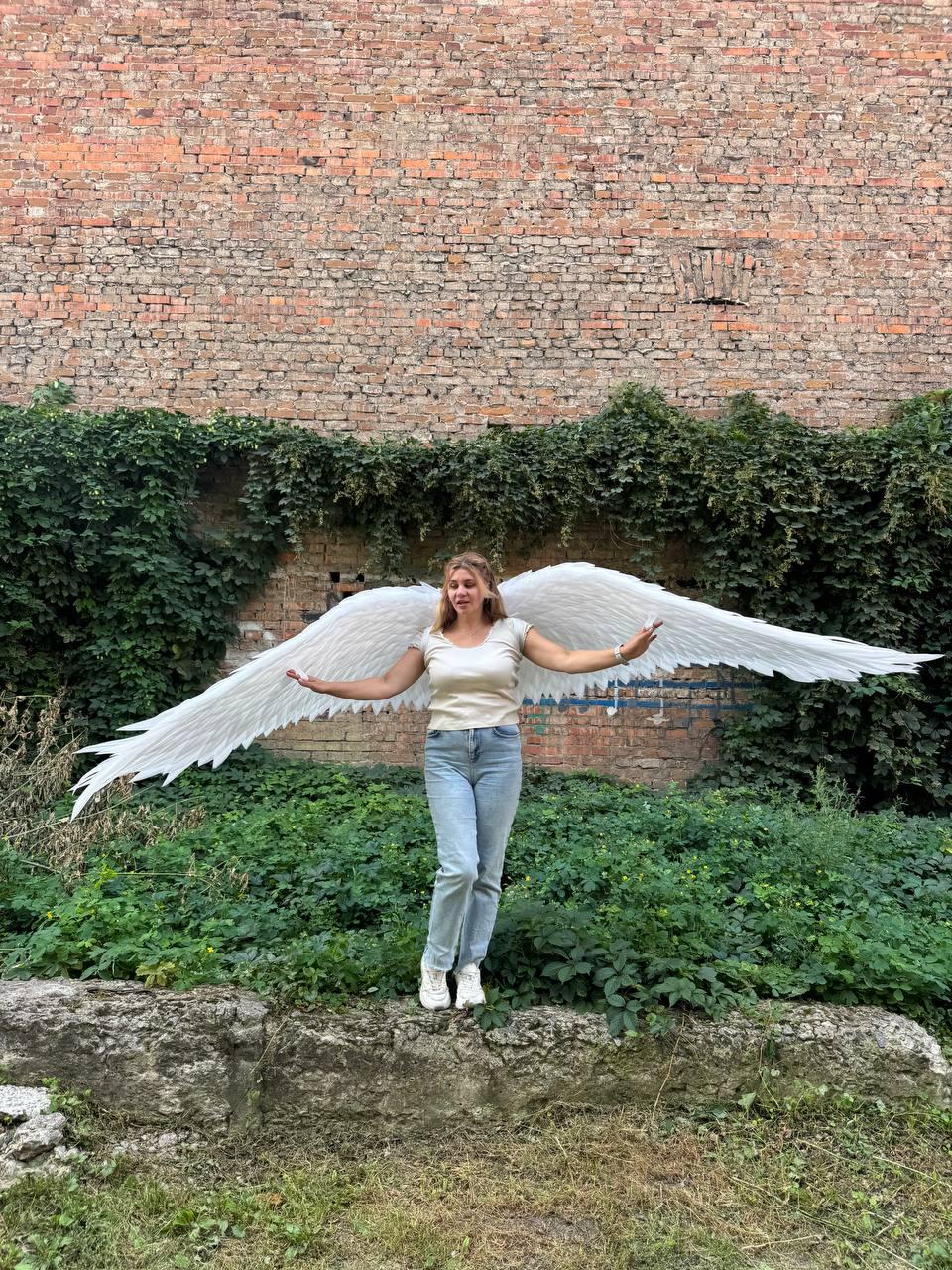 May include: A pair of large, white, feathered angel wings attached to a beige short-sleeved top and light blue denim jeans. The wings extend horizontally, featuring layered, pointed white feathers that span across the width of the frame.