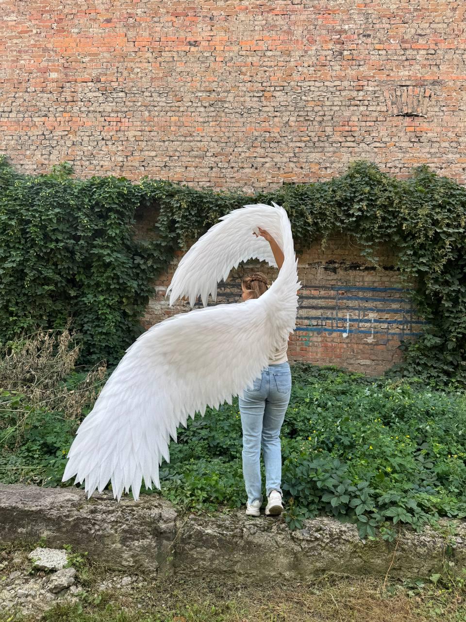 May include: A pair of large, white, feathered wings attached to a back harness. The wings feature layered, pointed white feathers that extend outward and upward, creating a wide, expansive span. The structure is designed to be worn on the back.
