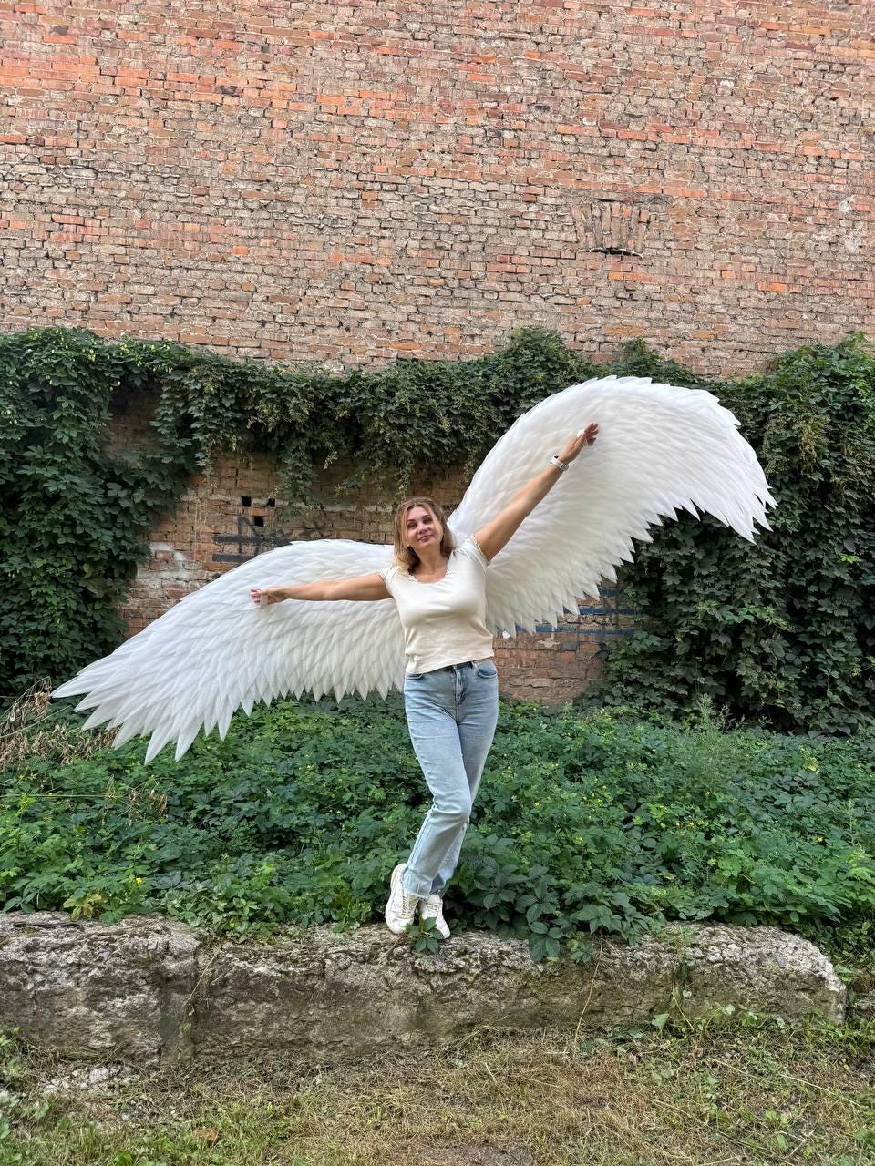 May include: A pair of large, white, feathered wings attached to a harness, worn by a person dressed in a cream-colored short-sleeved top and light blue denim jeans. The wings feature layered, pointed white feathers that extend outward in a wide span.