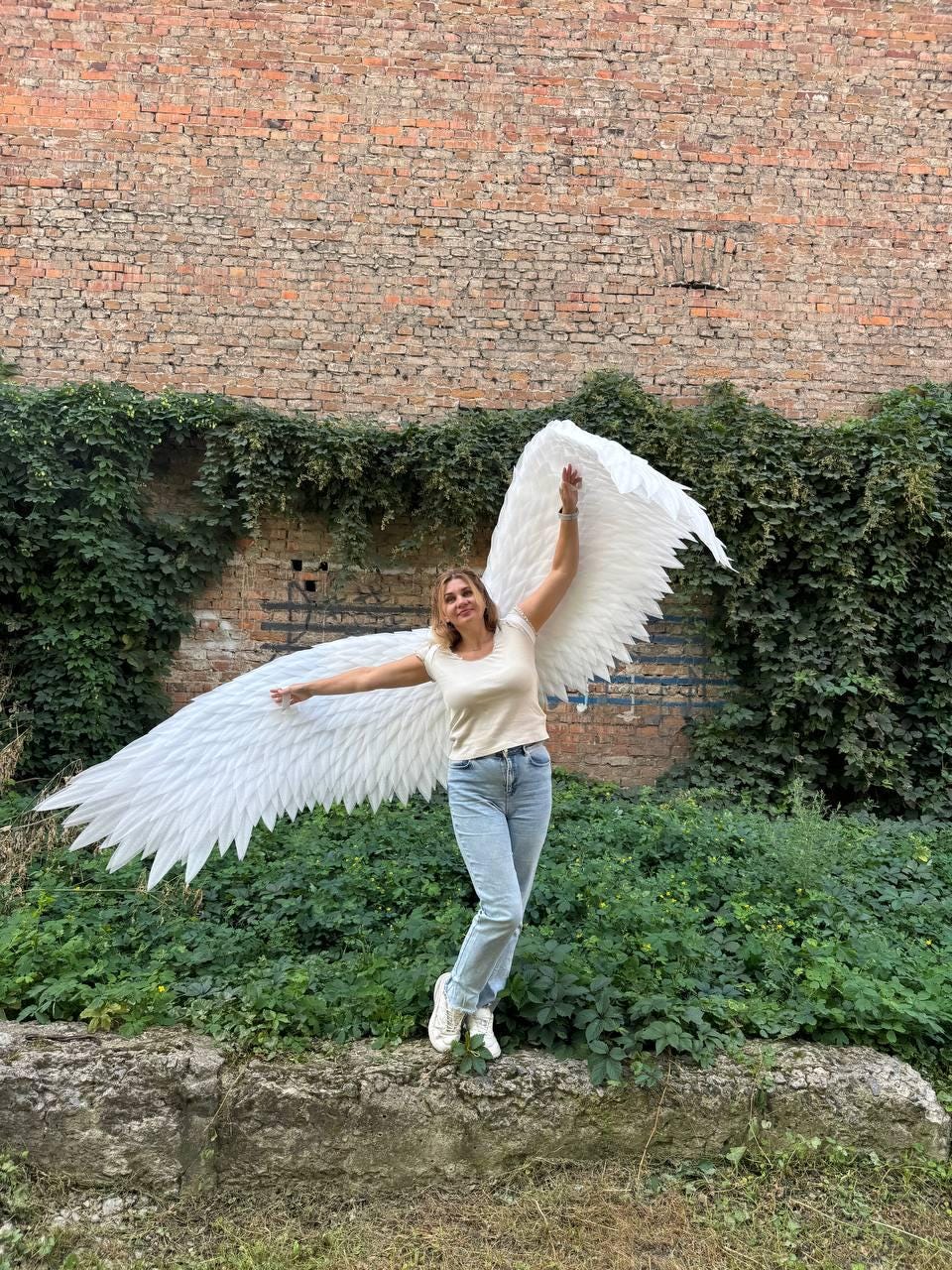 May include: Large white feathered wings attached to a beige short-sleeved t-shirt and light blue denim jeans. The wings feature layered, pointed white feathers that extend outward from the shoulders, creating a wide, expansive span.