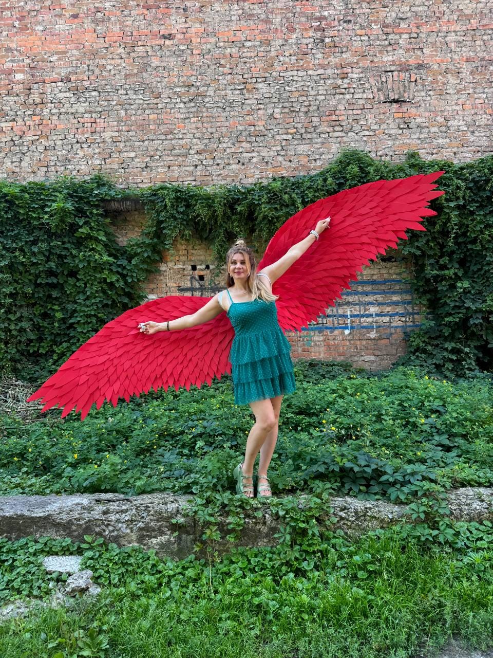 May include: A pair of large, vibrant red wings with layered feather-like segments attached to a teal, sleeveless, tiered mini dress. The wings extend horizontally, showcasing a wide span of bright red fabric shaped into pointed, overlapping feathers.
