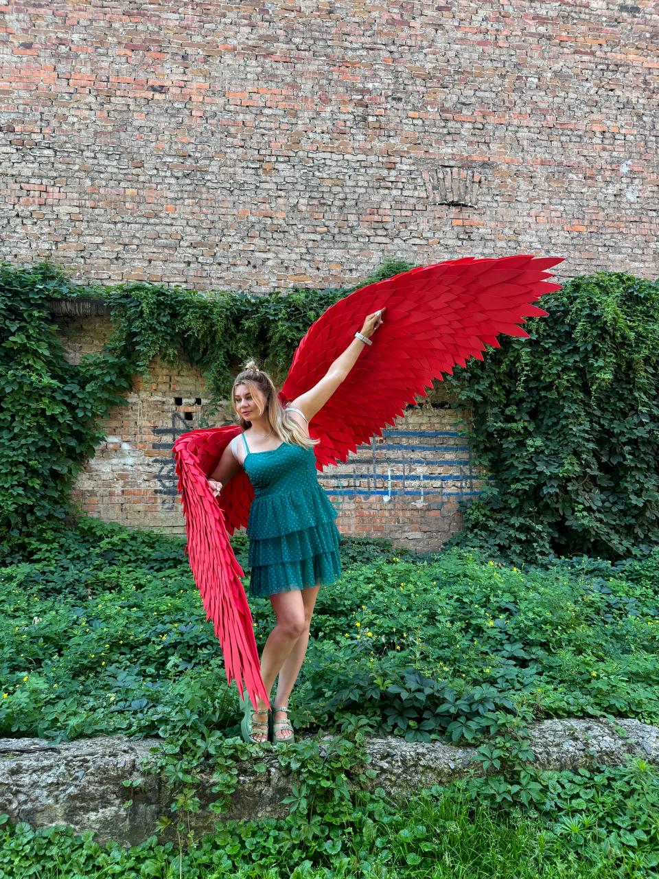 May include: A pair of large, vibrant red feathered wings attached to a back harness, worn with a sleeveless, knee-length, dark green tiered dress featuring a textured, dotted pattern and thin shoulder straps, paired with light green platform sandals.