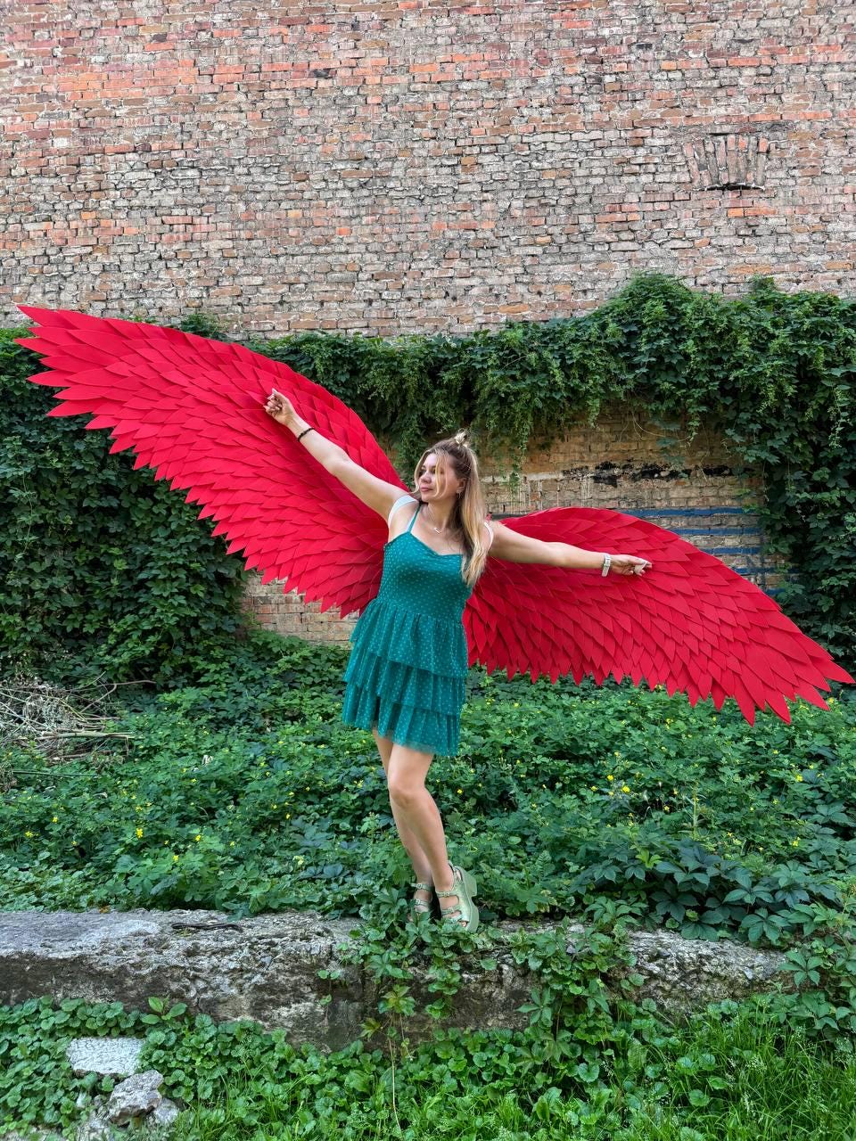 May include: A teal sleeveless dress with a tiered skirt and a textured pattern, paired with large, expansive, bright red feathered wings that extend horizontally, featuring layered, pointed feather-like segments across the entire span.
