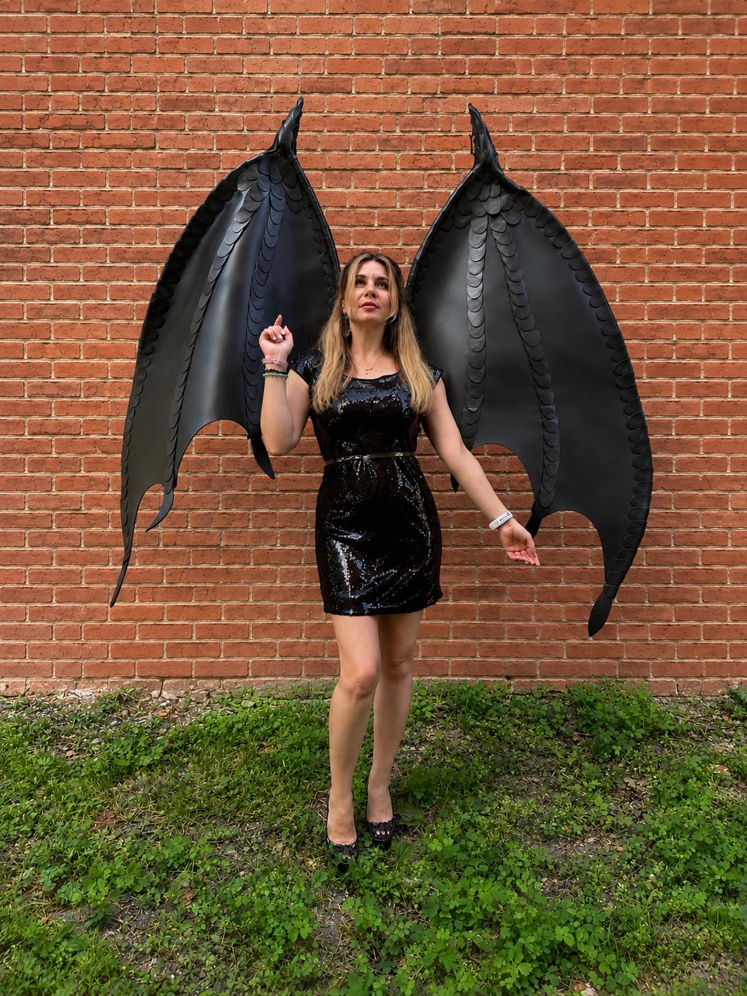May include: A black sequined mini dress paired with large, articulated black dragon wings featuring a textured, scale-like pattern along the upper edges. The wings extend upward and outward, showcasing a pointed, bat-like structure.
