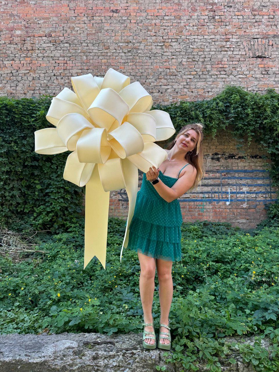 May include: A large, oversized cream-colored decorative bow with multiple loops and two long, hanging tails. The bow is constructed from a smooth, matte material and features a voluminous, rounded shape with a wide, structured ribbon design.