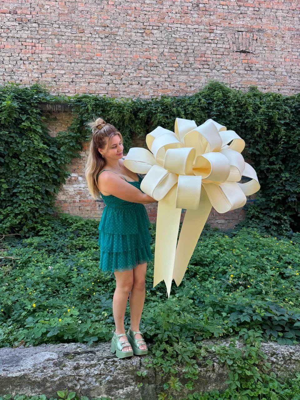 May include: A large, oversized cream-colored decorative bow with multiple loops and two long, wide ribbon tails featuring V-shaped cut ends. The bow is constructed from a smooth, matte material and is held in front of a person wearing a green dress.
