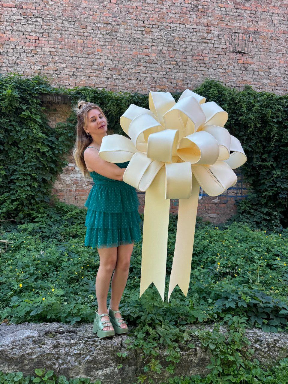 May include: A large, oversized cream-colored decorative bow with multiple rounded loops and two long, wide ribbon tails hanging downwards. The bow is constructed from a smooth, solid-colored material and features a voluminous, multi-layered design.