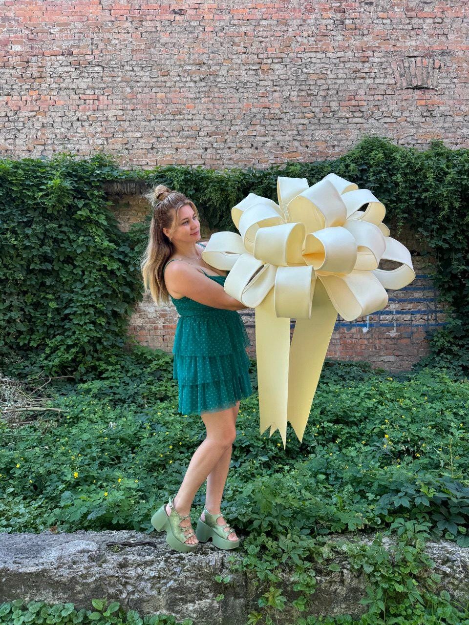 May include: A large, oversized cream-colored decorative bow with wide, smooth loops and two long, trailing ribbon tails. The bow is constructed from a stiff, matte material and features a complex, multi-layered design with a central knot.