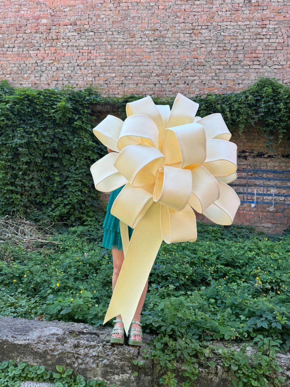 May include: A large, pale yellow decorative bow made of wide, smooth ribbon loops. The bow features a voluminous, multi-layered design with a single long, trailing ribbon tail extending downward from the center, creating a prominent focal point.