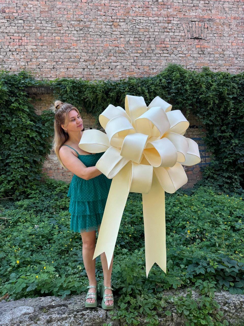 May include: A large, oversized decorative bow in a pale cream color, featuring multiple voluminous loops and two long, wide ribbon tails with V-shaped cut ends. The bow is constructed from a smooth, solid-colored material with a subtle sheen.