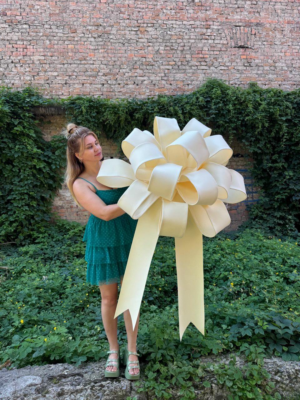 May include: A large, oversized decorative bow in a pale cream color, featuring multiple voluminous loops and two long, wide ribbon tails with V-shaped cut ends. The bow is constructed from a smooth, solid-colored material with a subtle sheen.