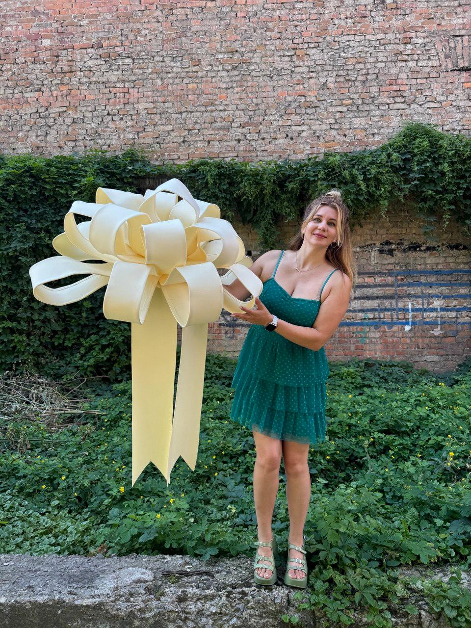May include: A large, oversized decorative bow in a pale cream color, featuring multiple wide, structured loops and two long, hanging ribbon tails. The bow is constructed from a smooth, matte material and is held up by a person.