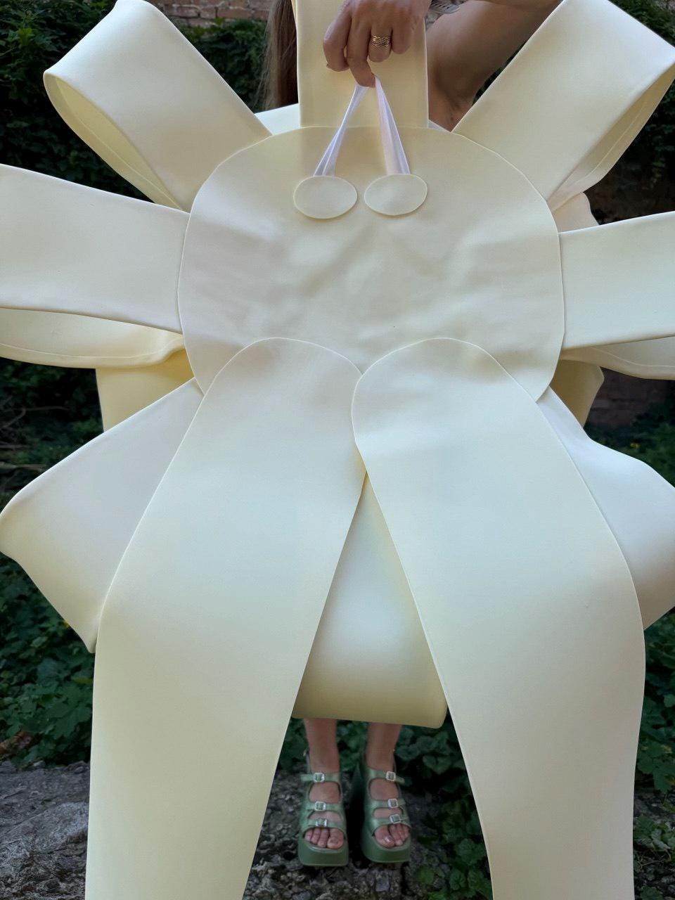 May include: A large, pale yellow, oversized bow-shaped accessory constructed from smooth, matte material. The design features a central circular panel with two small, circular accents at the top, connected by thin white straps, and multiple wide, radiating loops.