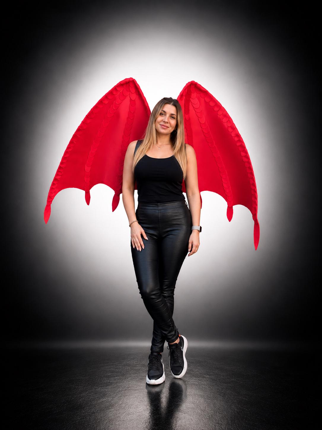 May include: Large, vibrant red fabric wings with a structured, pointed design and visible stitching details. The wings feature a bat-like shape with multiple downward-pointing tips, extending outward from the back of the wearer.