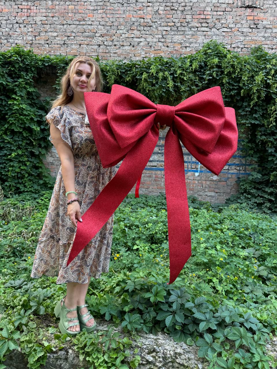 May include: A large, oversized decorative bow in a deep red color with a subtle glittery texture. The bow features two wide, voluminous loops and two long, trailing ribbon tails that hang vertically, creating a prominent and festive appearance.