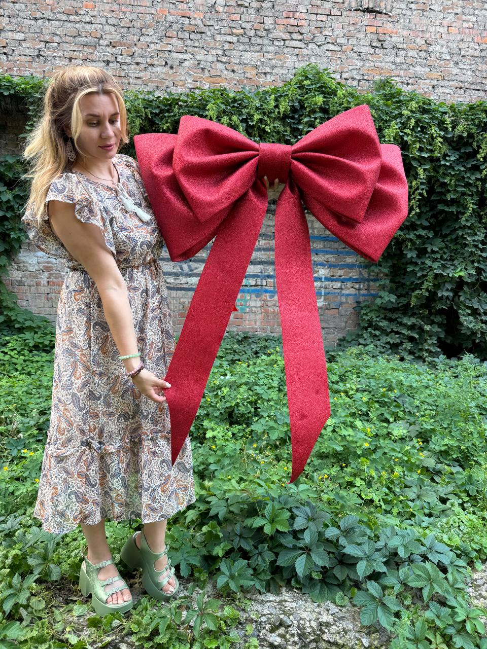 May include: A large, oversized decorative bow in a vibrant, glittery red finish. The bow features two wide, structured loops and two long, trailing ribbon tails that extend downward. The surface has a distinct sparkling texture throughout.