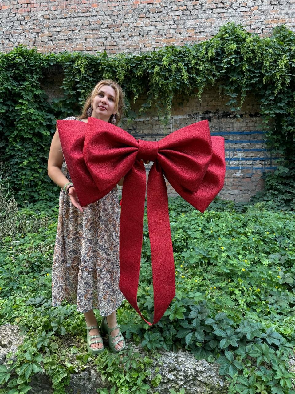 May include: A large, oversized decorative bow in a deep red color with a glittery, textured finish. The bow features two wide, prominent loops and two long, hanging ribbon tails that extend downward, creating a bold and festive appearance.