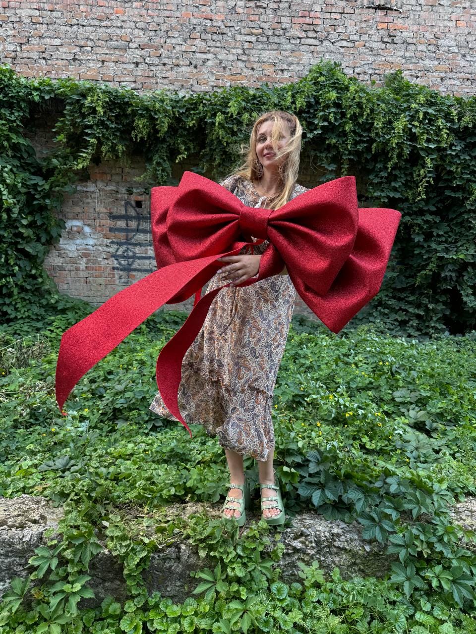May include: A large, oversized red bow with a textured, glittery finish. The bow features two prominent loops and two long, trailing ribbon ends that drape downwards. The material appears stiff and structured, maintaining a wide, expansive shape.