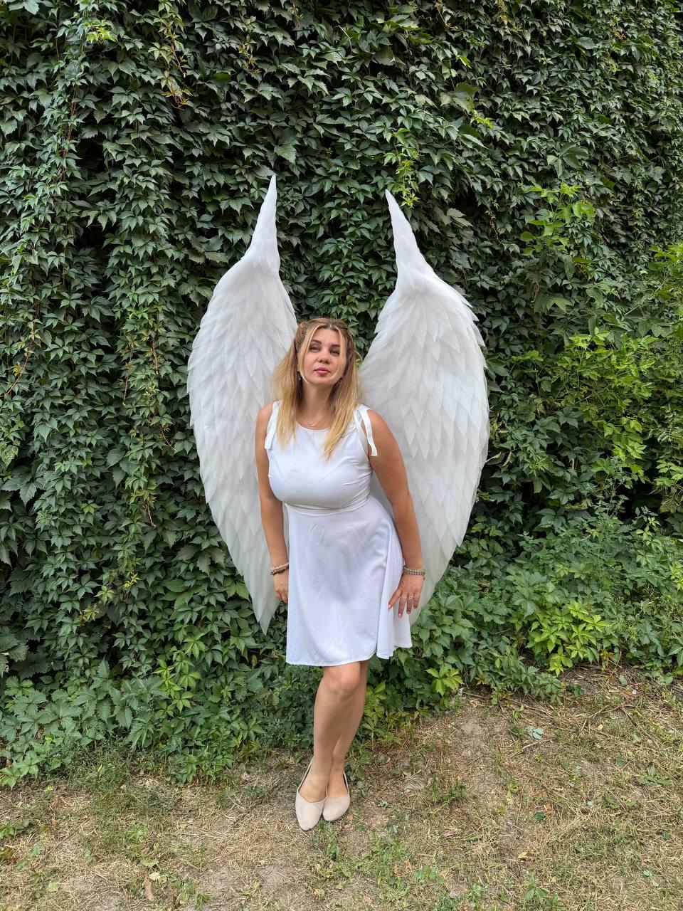 May include: A pair of large, white, feathered angel wings attached to a white sleeveless dress. The wings are tall, pointed at the top, and feature layered, textured plumage. The dress is a simple, solid white knee-length garment with thin straps.