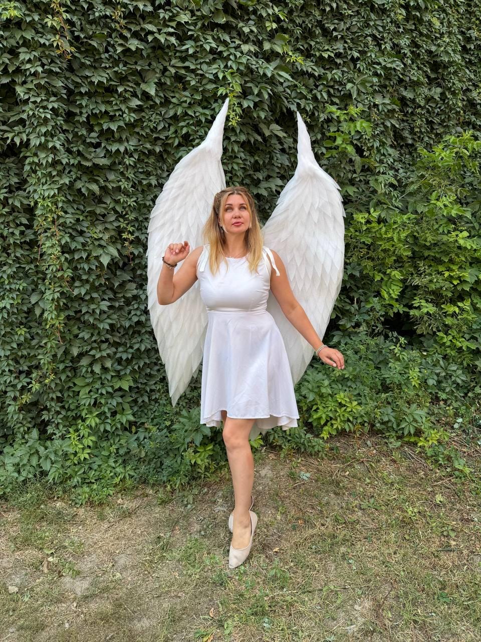 May include: A white sleeveless dress with a flared skirt and shoulder ties, paired with large, tall, white feathered angel wings. The wings feature layered, pointed feathers that extend upward and outward, creating a dramatic, expansive silhouette.