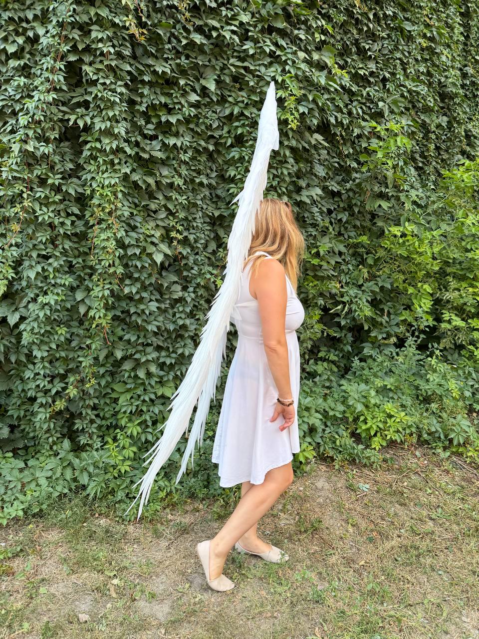 May include: A sleeveless white dress paired with a large, tall set of white feathered wings that extend vertically from the back. The wings feature long, slender, layered white feathers that taper to a point at the top and bottom.