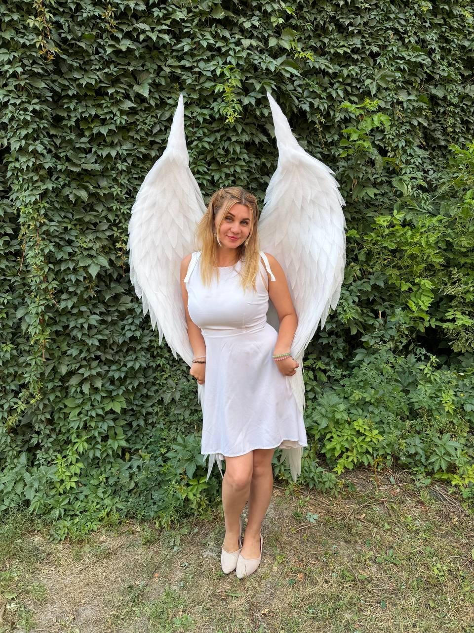 May include: A white sleeveless dress with a round neckline and a flared skirt, paired with large, tall, white feathered wings that extend upwards behind the shoulders. The wings feature layered, pointed white feathers in a structured design.