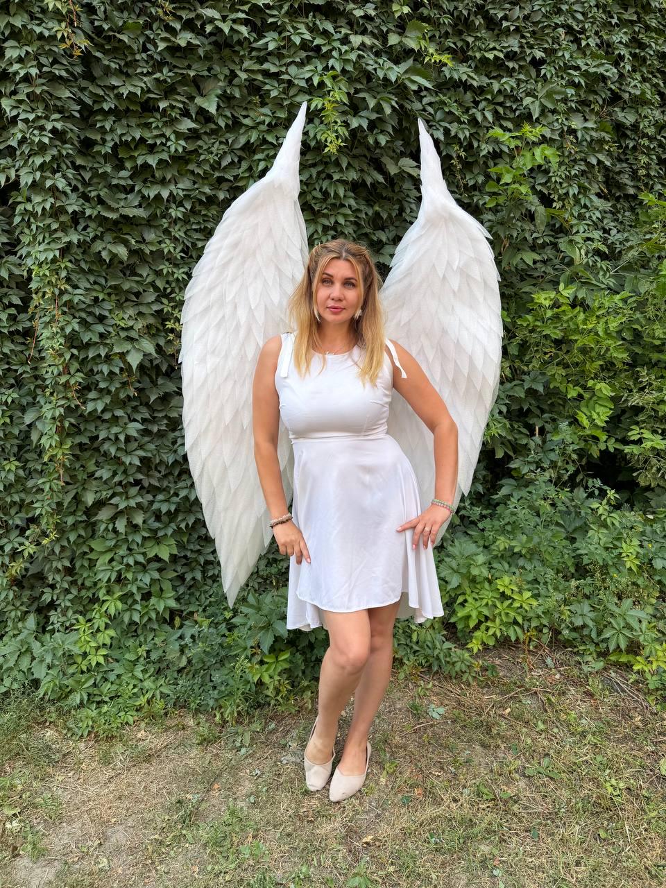 May include: A white sleeveless dress with a scoop neckline and a flared skirt, paired with large, tall, white feathered wings that extend upwards behind the shoulders, featuring layered plumage and a pointed, arched shape at the top.