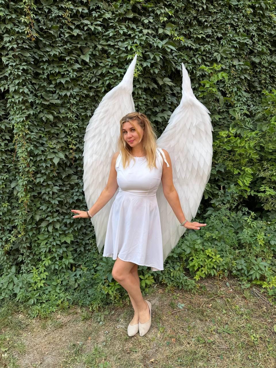 May include: A white sleeveless dress with a flared skirt paired with large, feathered white angel wings. The wings are tall, pointed, and textured with layered white feathers, extending upwards behind the shoulders of the white dress.