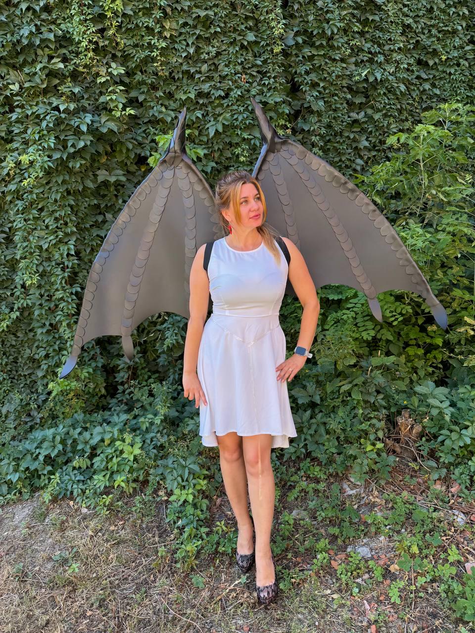 May include: A white sleeveless dress with a fitted bodice and a flared skirt, paired with large, dark gray, textured dragon-style wings worn on the back with black straps. The wings feature a scalloped edge design and a rigid, structured appearance.