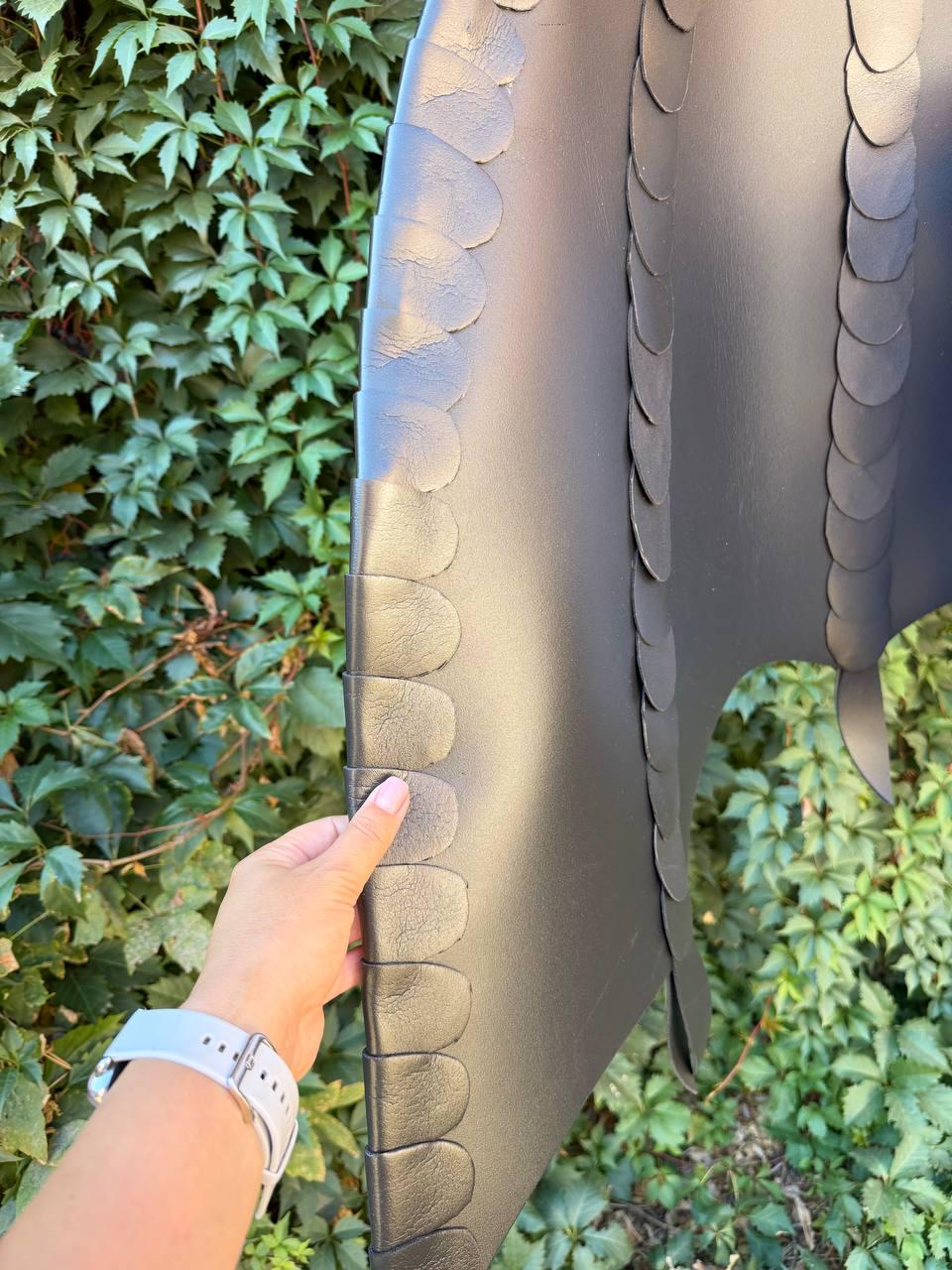 May include: A black leather cape featuring vertical rows of overlapping, rounded scale-like cutouts. The garment is constructed from smooth, matte black material with distinct, layered edges that create a textured, dimensional appearance.