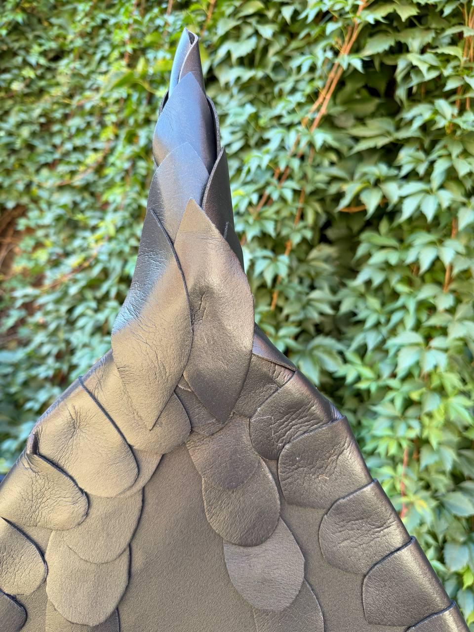 May include: A dark gray leather accessory featuring a pointed, conical shape constructed from overlapping, petal-like leather segments. The surface displays a natural grain texture, with the layered pieces creating a scale-like pattern.