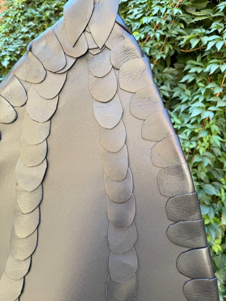 May include: A black leather accessory featuring a triangular shape adorned with multiple rows of overlapping, petal-shaped leather cutouts arranged in vertical lines. The surface has a smooth texture, and the edges are finished with these distinct, layered leather scales.