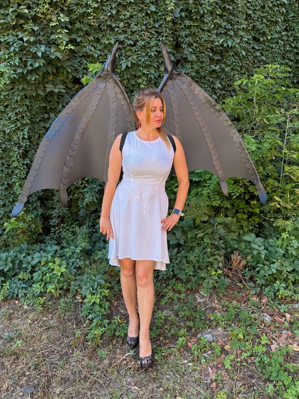 May include: A white sleeveless knee-length dress with a simple round neckline and a thin silver-toned chain belt at the waist, paired with large, dark gray, textured dragon-style wings featuring a scalloped edge and black shoulder straps.
