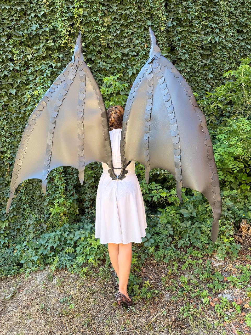May include: Large, matte black dragon wings featuring a structured, layered scale design with pointed tips and a central harness mechanism. The wings are expansive, extending upward and outward with a rigid frame and distinct, segmented wing panels.