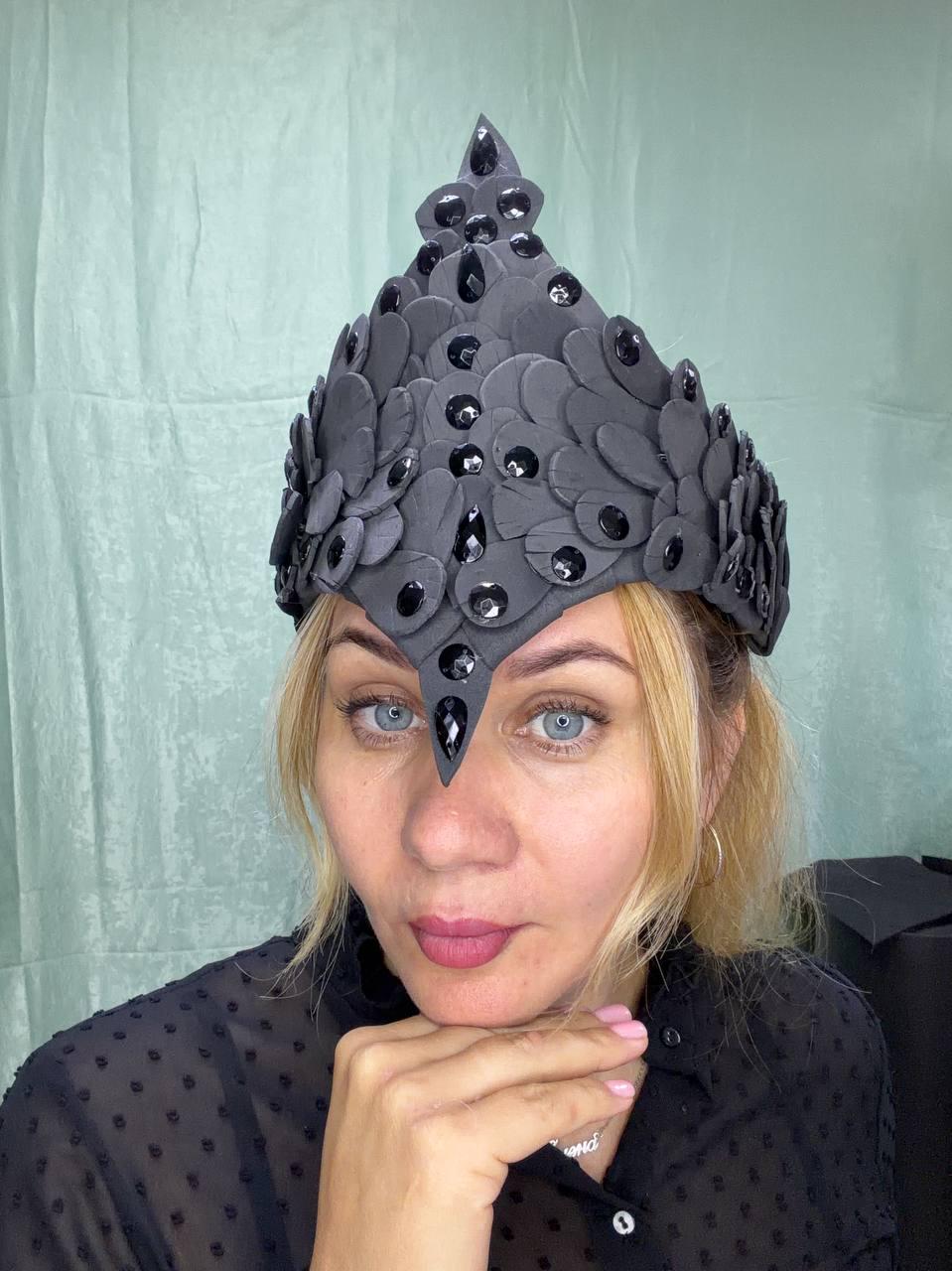 May include: A dark gray headpiece featuring a pointed, crown-like structure constructed from layered, petal-shaped pieces. The surface is adorned with numerous black, faceted, teardrop-shaped crystals arranged in a symmetrical pattern.
