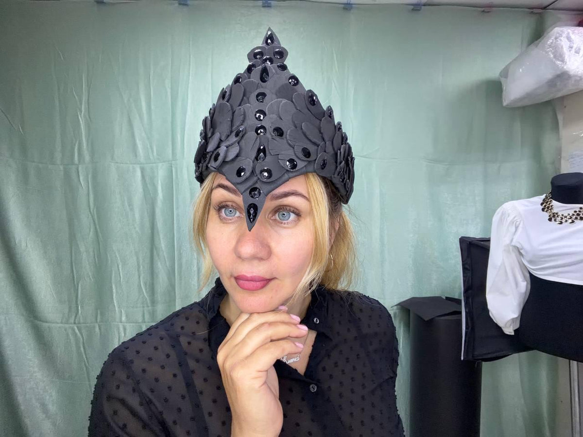 May include: A pointed black headpiece featuring layered petal-like shapes and embedded dark, glossy, teardrop-shaped stones. The structure rises to a central peak, with the decorative elements arranged in a symmetrical pattern across the front.