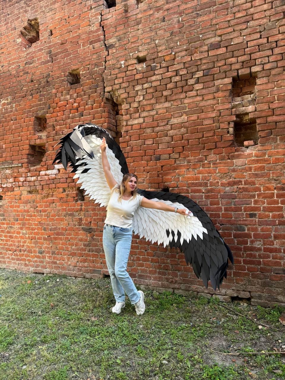 May include: A pair of large, wearable wings featuring layered black and white feathers. The wings are constructed with a structured design, displaying a gradient of white feathers near the base transitioning to black feathers at the outer edges.