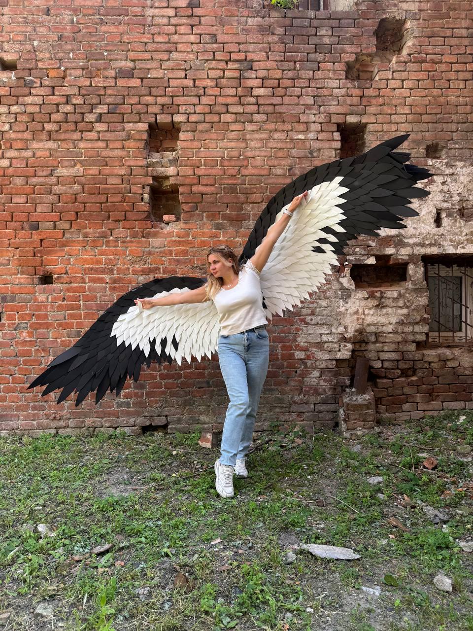 May include: A pair of large, wearable wings featuring a layered design with white inner feathers and black outer feathers. The wings are constructed with a rigid structure and are attached to a harness, displaying a wide, expansive wingspan.